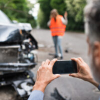 Image of a man taking a picture of a crashed car, representing the need to document details of a hit and run accident and contact a WV hit and run lawyer like those at Burke, Schultz, Harman & Jenkinson as soon as possible.